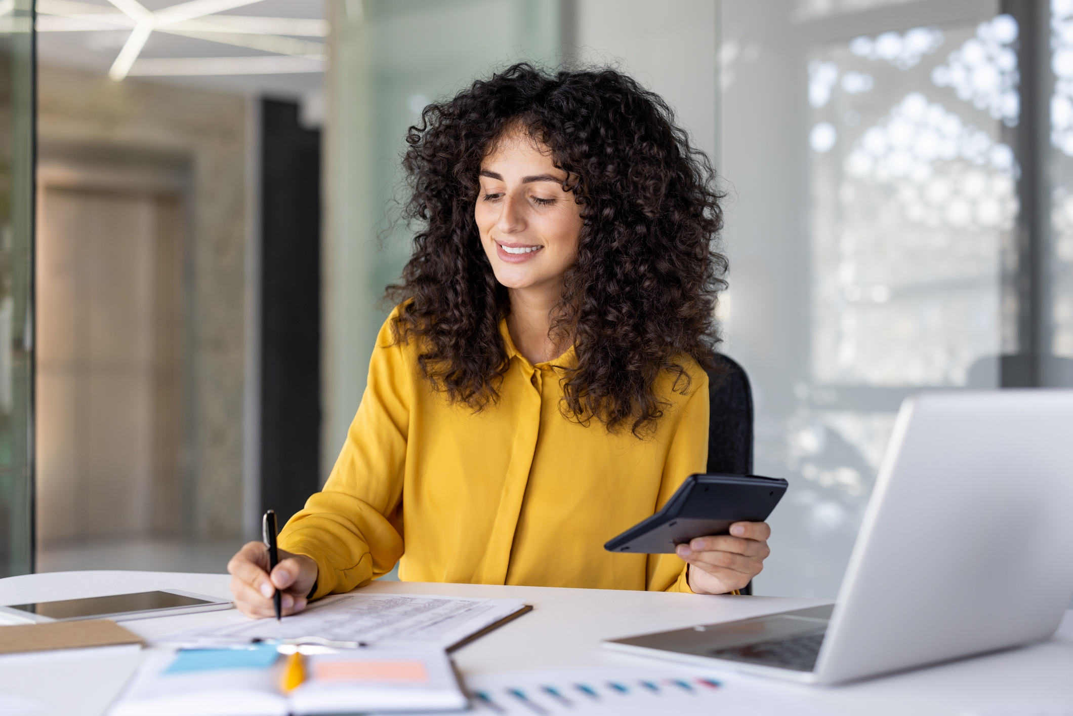 Successful satisfied female financier doing paperwork inside office. Businesswoman holding calculator in hands and smiling writing down filling out tax forms and investment reports.