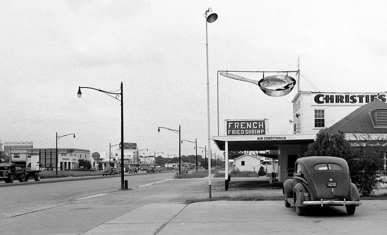 A black-and-white photo of the exterior of Christie's Seafood and Steaks in Houston, Texas, next to a road. An old-fashioned car is in Christie's parking lot.