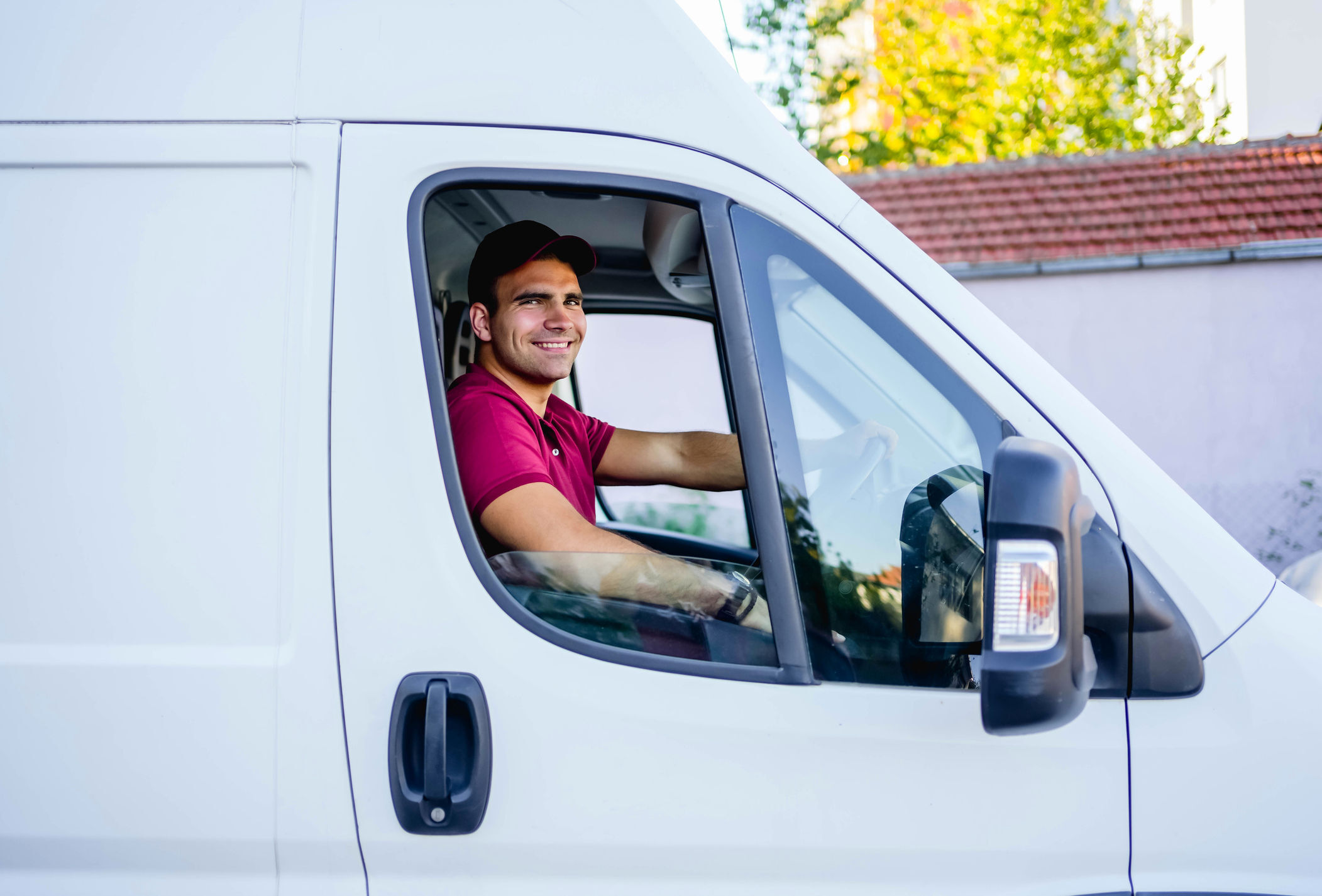 A man is seated in the driver's seat of a delivery van. He is wearing a hat and smiling at the viewer. He is in the process of making deliveries for his employer.