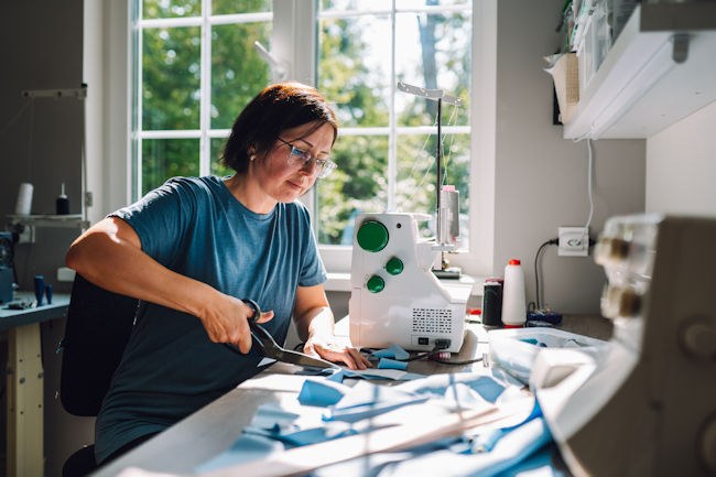 A woman is seated before a sewing machine in a sunlit room. She is holding a pair of scissors and cutting fabric.
