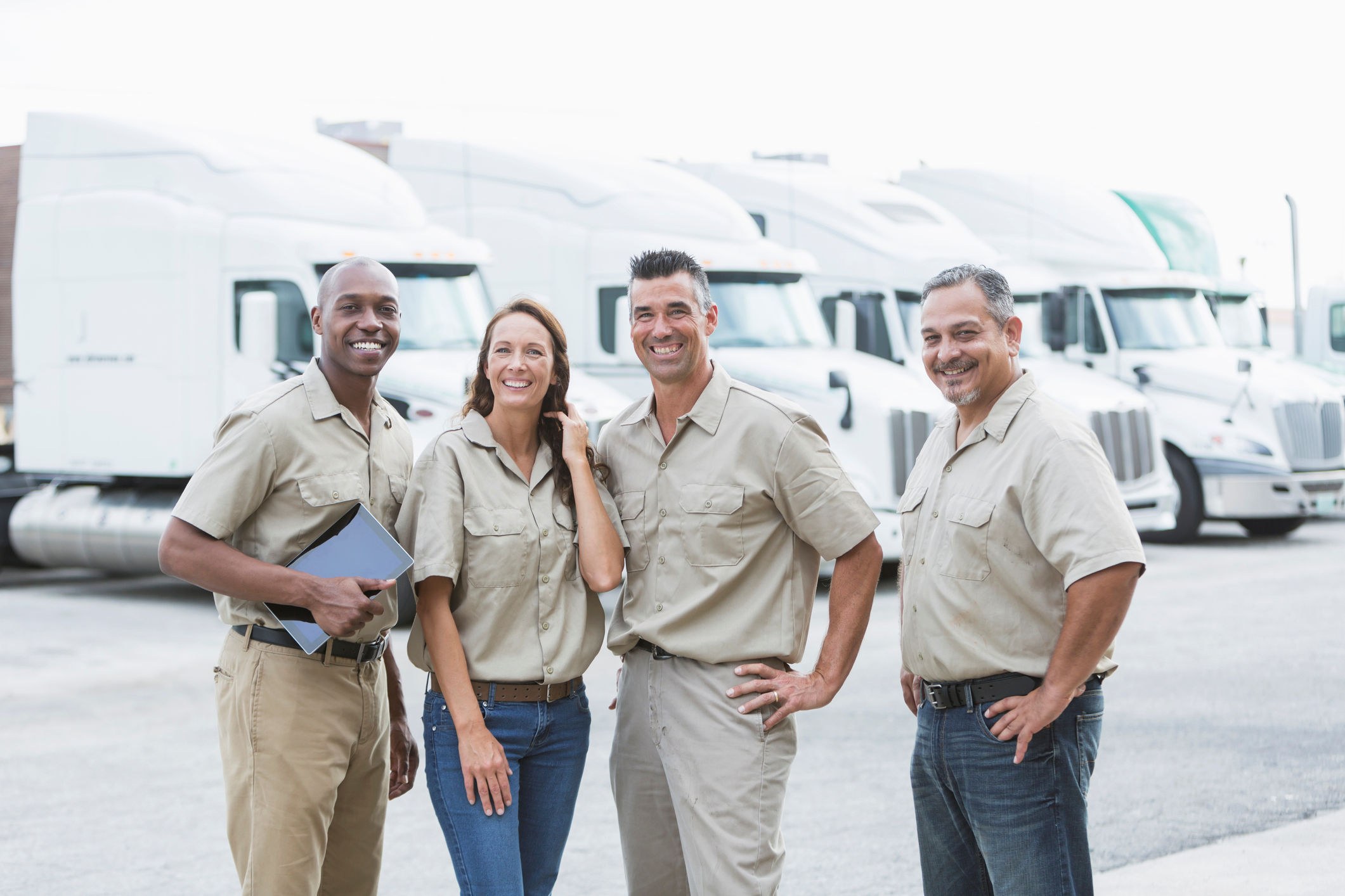 A group of company workers stand in front of a fleet of semitrucks that are parked at a warehouse distribution facility.