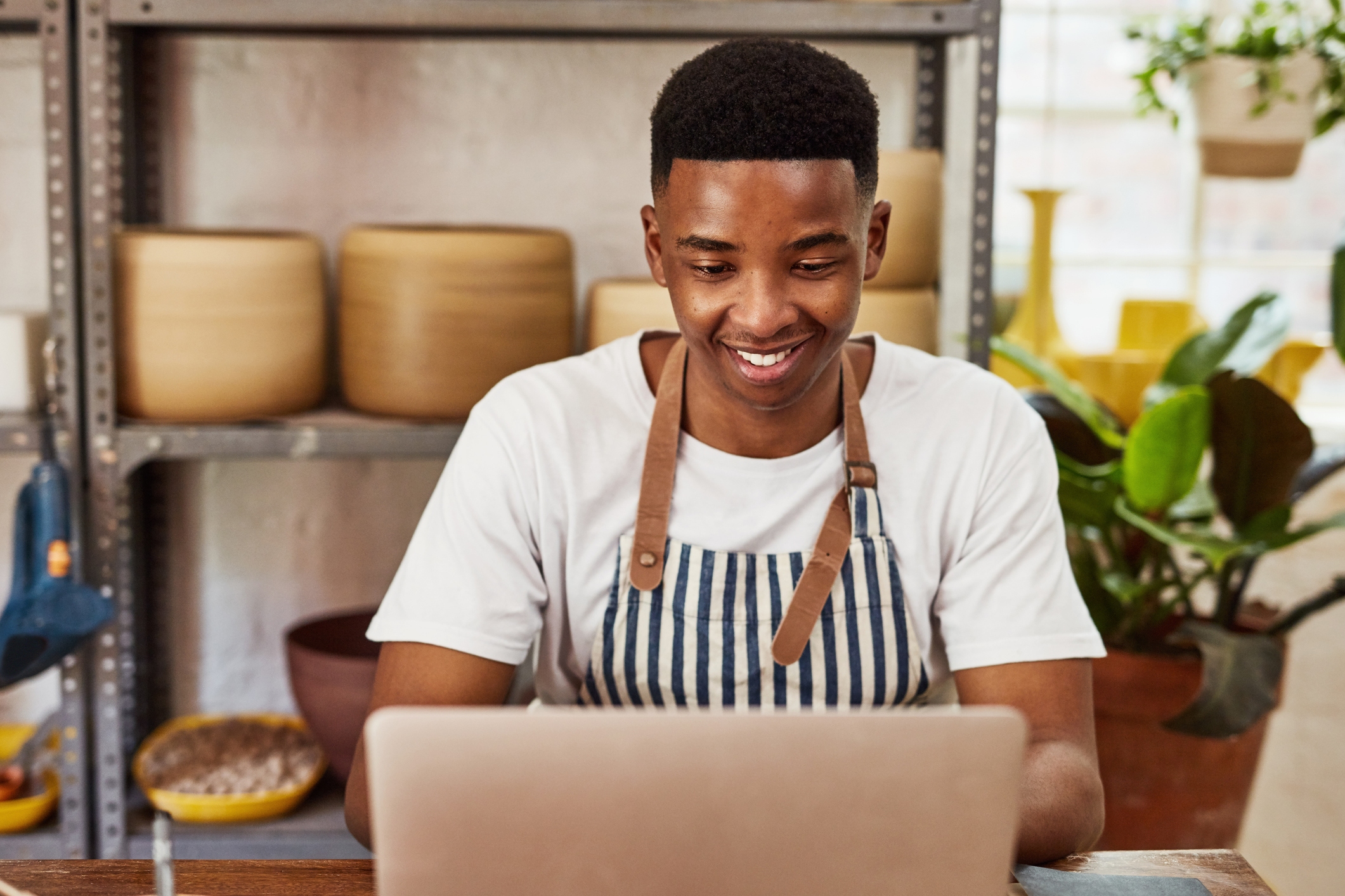 A young man sits at a laptop smiling. He wears a white T-shirt under a blue-and-white striped apron. Behind him are shelving units filled with large ceramic pots.