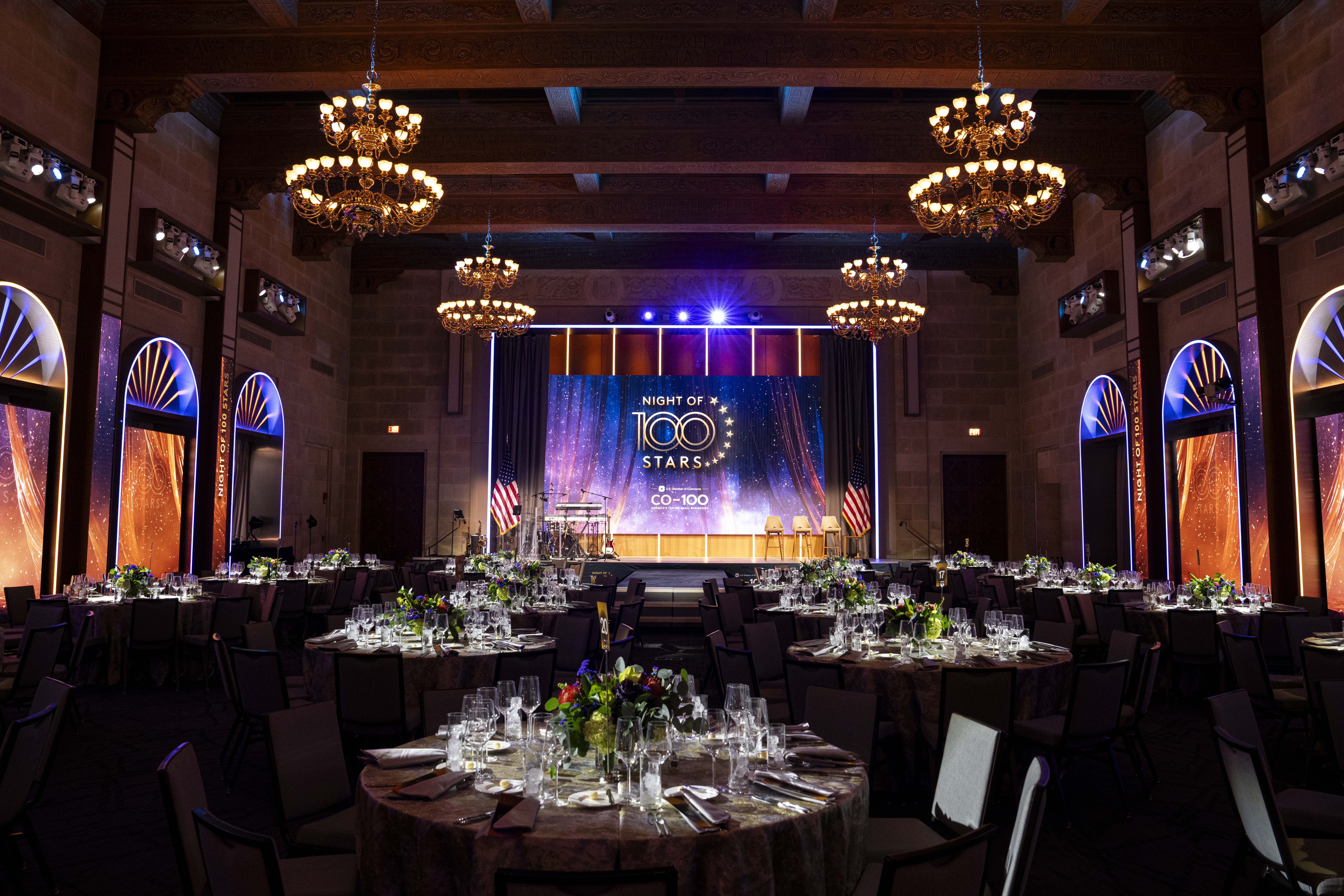 An elegant banquet hall with round tables set for a formal event, illuminated by ornate chandeliers and a stage displaying "Night of 100 Stars" with American flags.