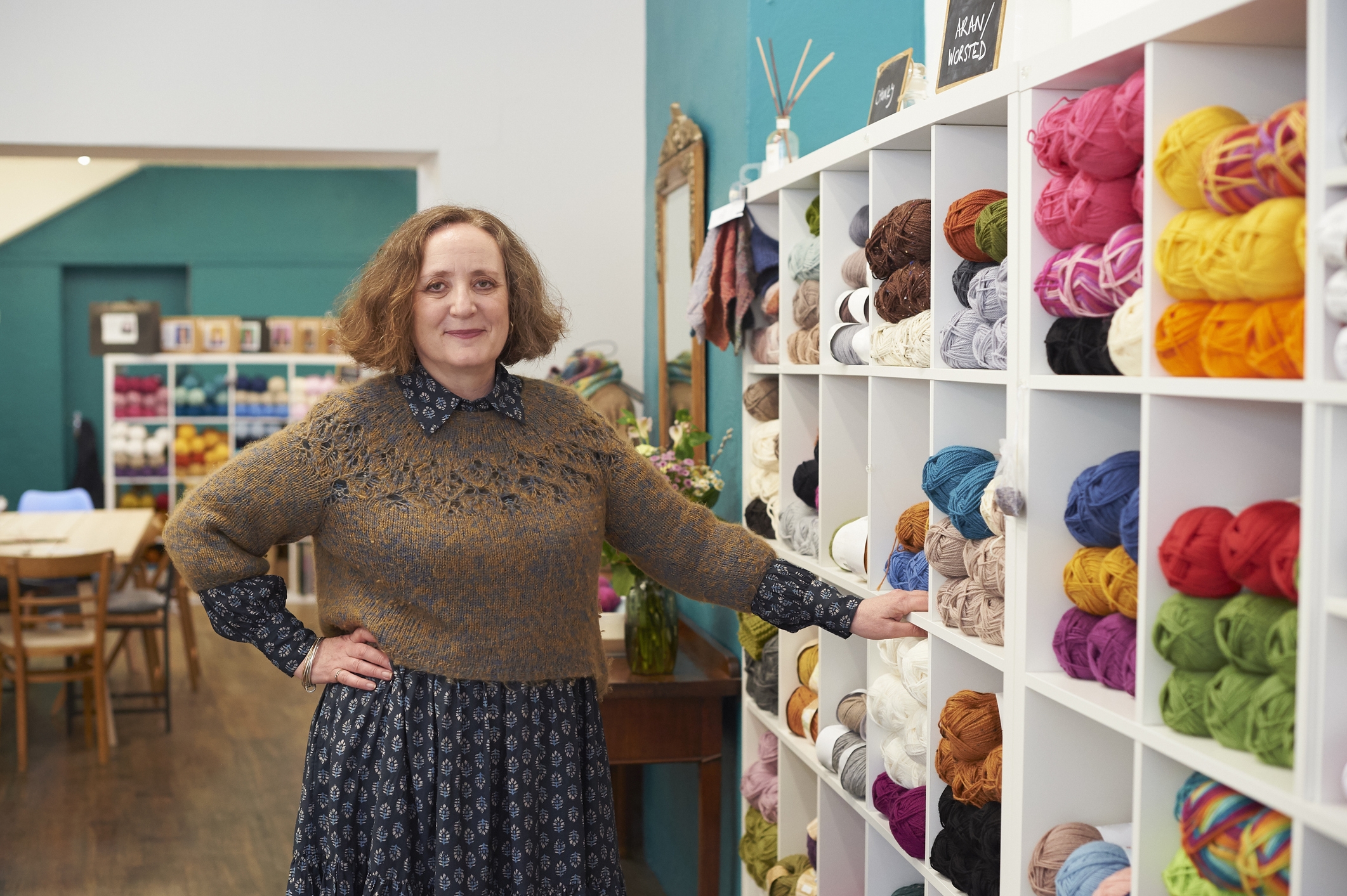 The owner of a yarn store, a middle-aged woman, stands next to cubbyhole shelves of yarn in various colors. The store owner has bobbed auburn hair and she wears a blue patterned dress under a brown sweater. She poses with one hand on the shelves and one on her hip.