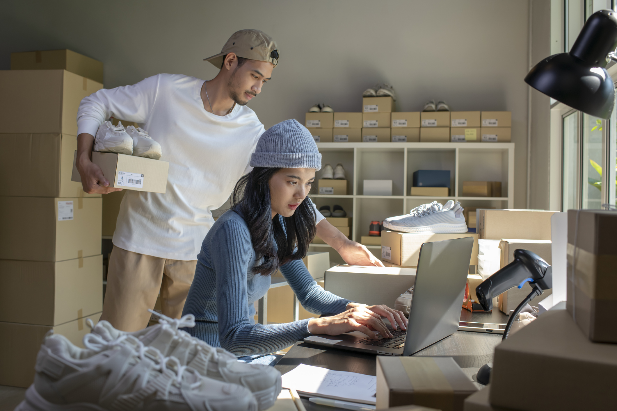 A woman and a man work in a small storeroom filled with stacked cardboard boxes and shelves of shoe boxes. The woman sits at a desk and works on a laptop; she has long dark hair and wears a long-sleeved blue shirt and a light blue beanie. The man is standing behind her, moving boxes. He holds a shoebox and a pair of white sneakers under one arm as he reaches for another box. The man has dark hair and a goatee, and he wears a white long-sleeved shirt, khakis, and a gray backwards baseball cap.