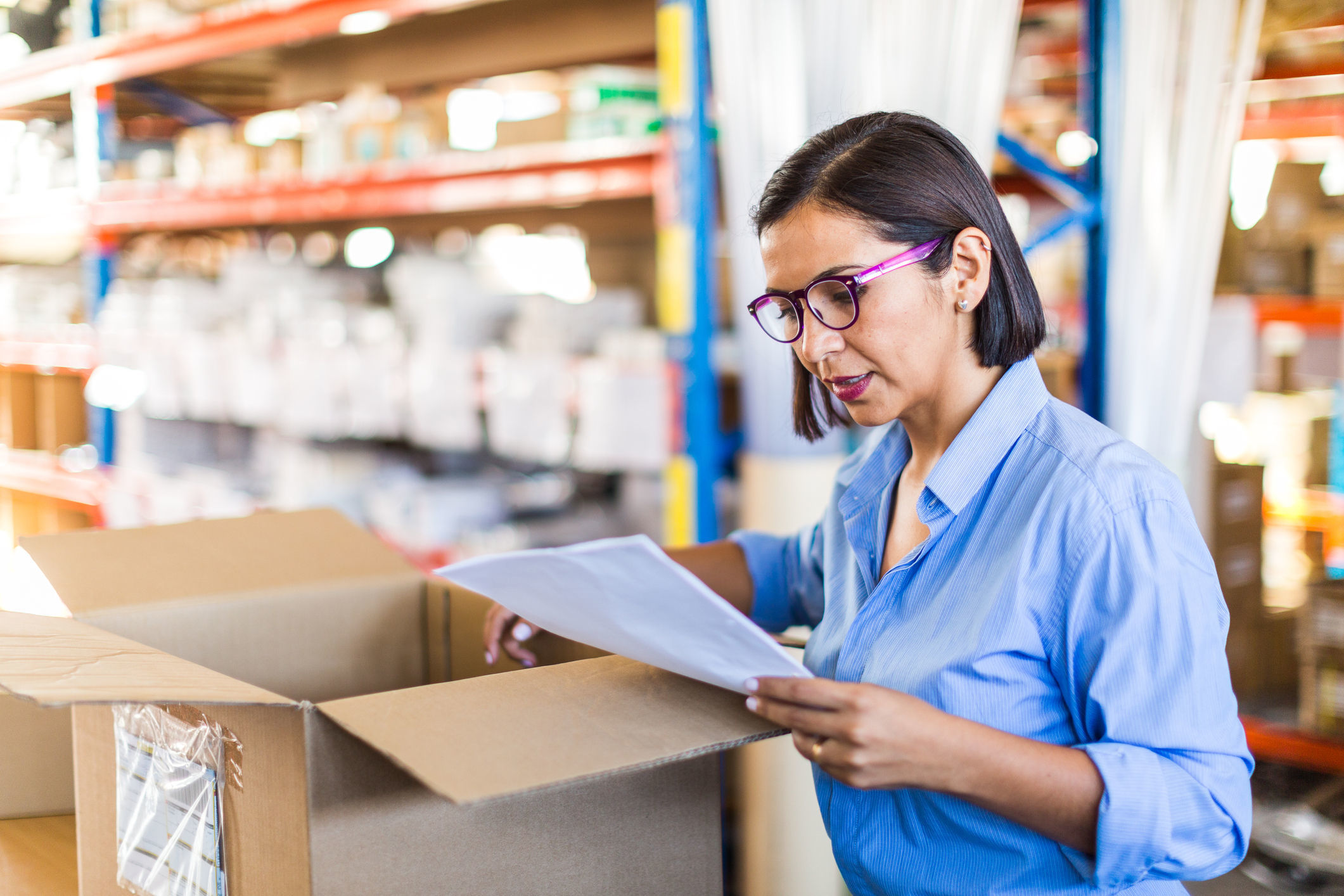 A small business owner checks a delivery. Before her is an open box. She is holding the shipping invoice in her right hand and comparing it against the goods delivered.