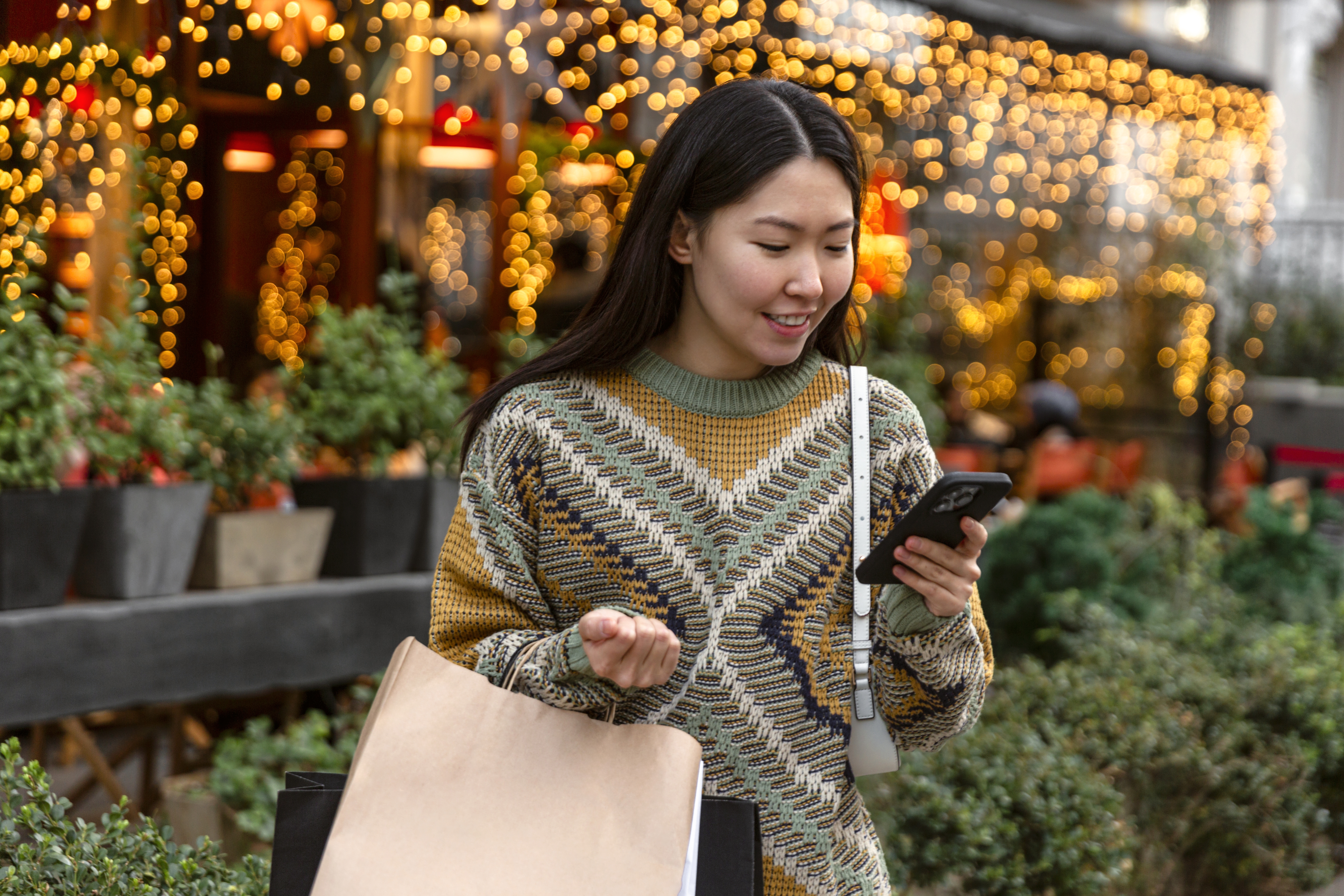 A woman holding shopping bags looks down at her smartphone with a smile. In the background are lines of potted bushes and small fir trees under an awning hung with dangling lights.