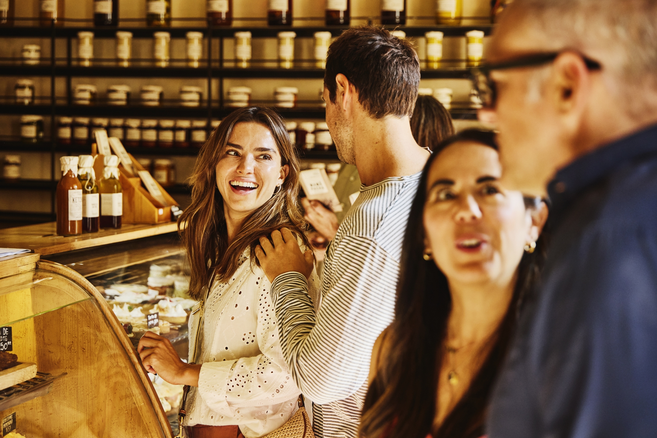 A group of people standing in a bakery, crowded near the display cases, talking to each other and smiling. The light in the room is golden and the walls are lined with shelves of jars in various sizes.