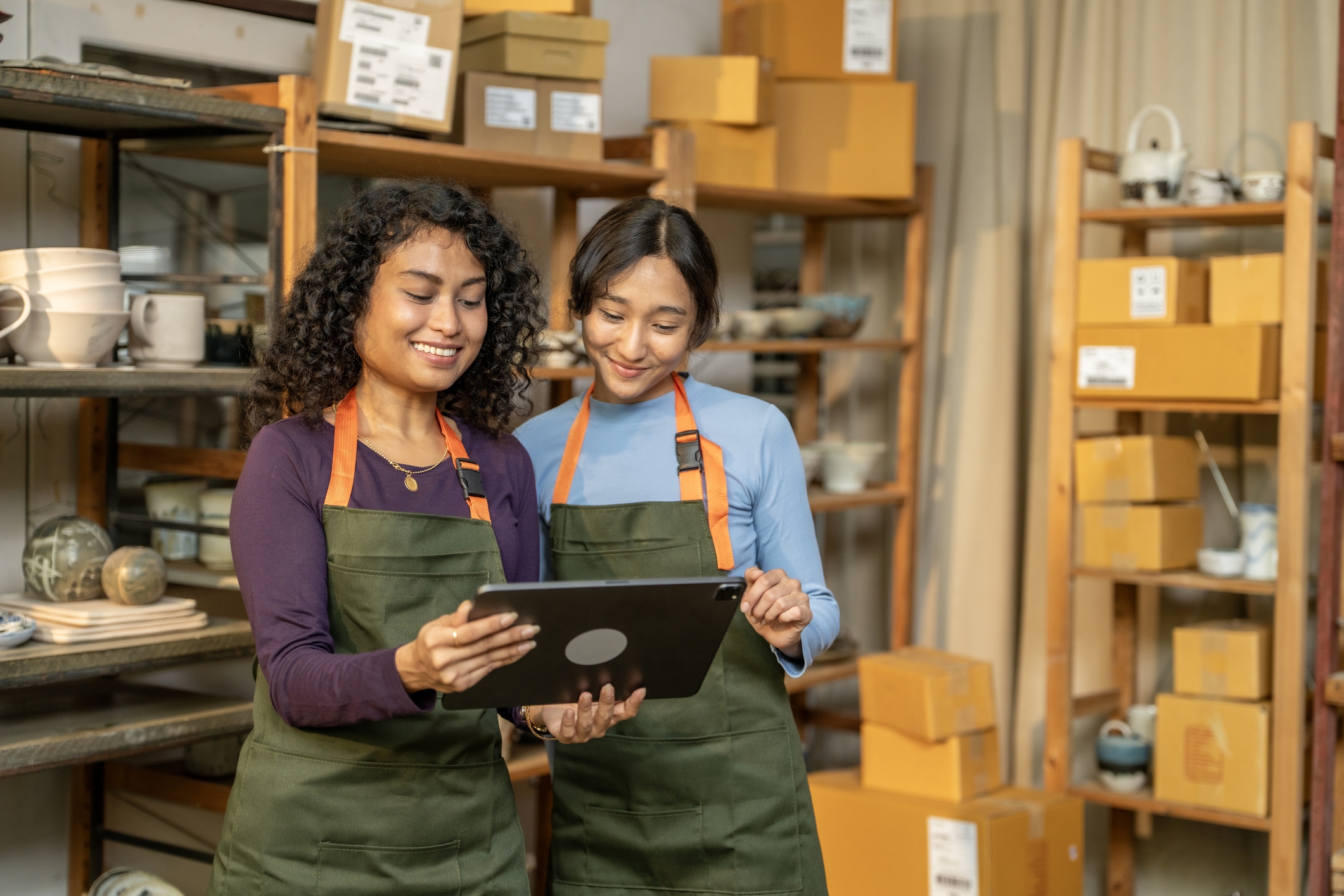 Two women wearing aprons are standing next to each other in a storage room, looking at a tablet. In the background are shelves of cardboard boxes and ceramic dishes and mugs.