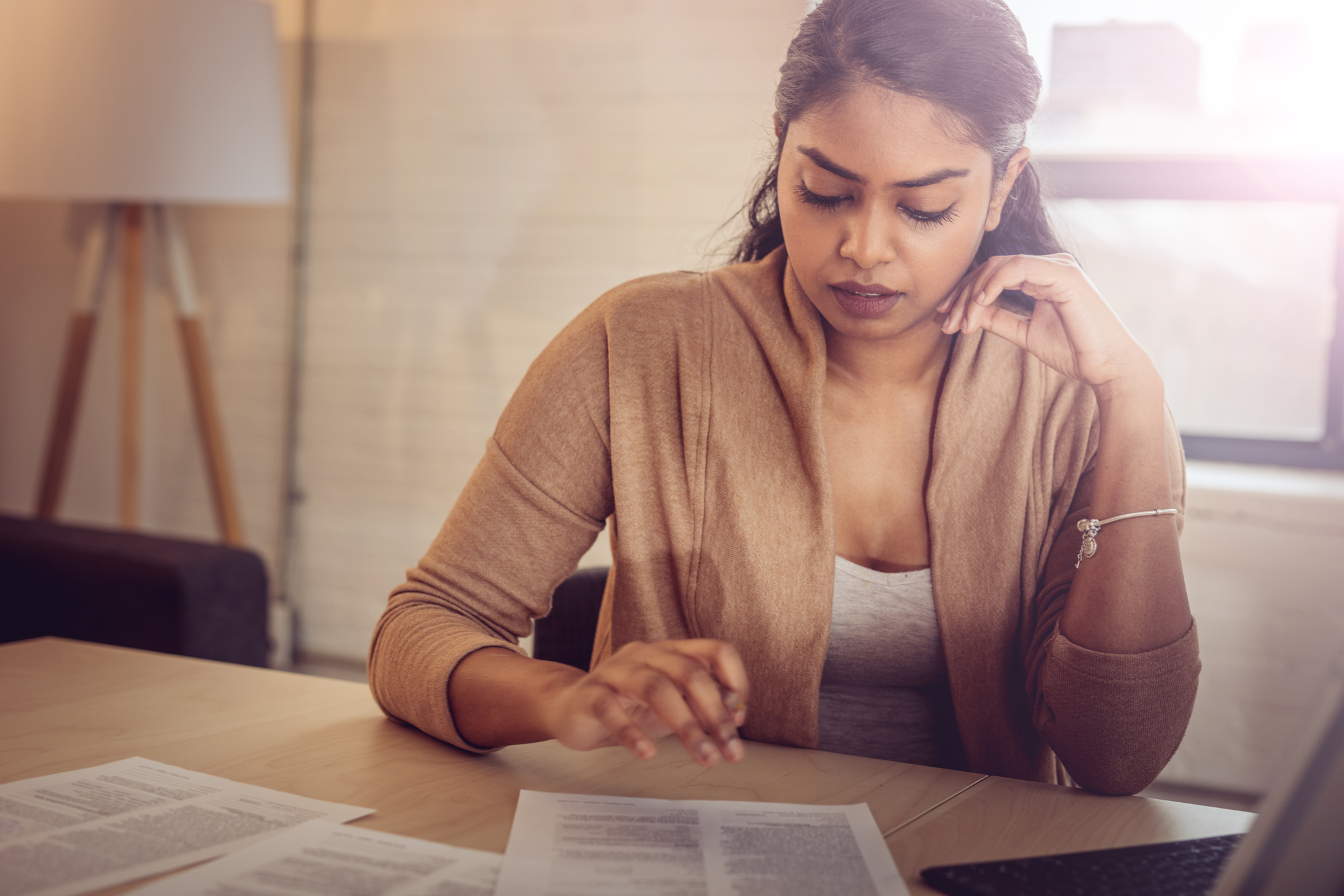 A woman sits at a table and looks down thoughtfully at a few scattered papers.
