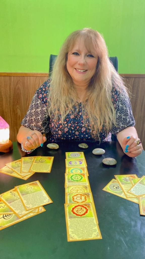 Descubre tu guía interior y equilibrio de chakras a través de oráculos y cartas florales Instructora sonriente con cartas de oráculo de chakras y piedras en un escritorio, ofreciendo guía espiritual y equilibrio energético en Mago Holístico