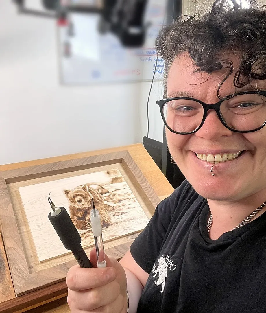 Pyrography artist Kez Halliday smiling with her pyrography pens and a framed bear portrait Pyrography artist Kez Halliday smiling with her pyrography pens and a framed bear portrait