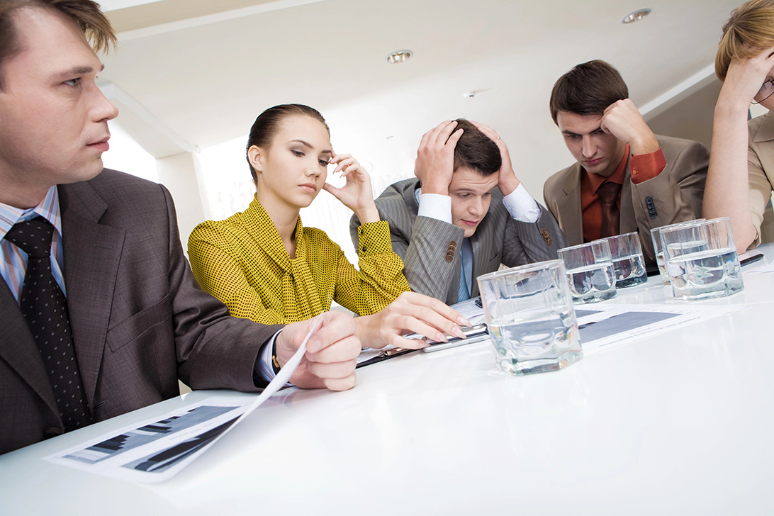 Grupp med affärspersoner som sitter vid mötesbord och ser trötta och frustrerade ut, symbol för brist på uppskattning och negativ arbetsmiljö.