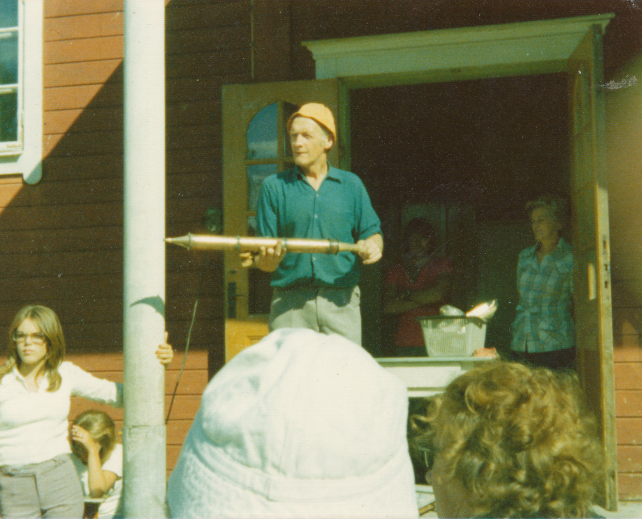 Färgfotografi av Emil stående utanför en byggnad, omgiven av människor under en auktion.