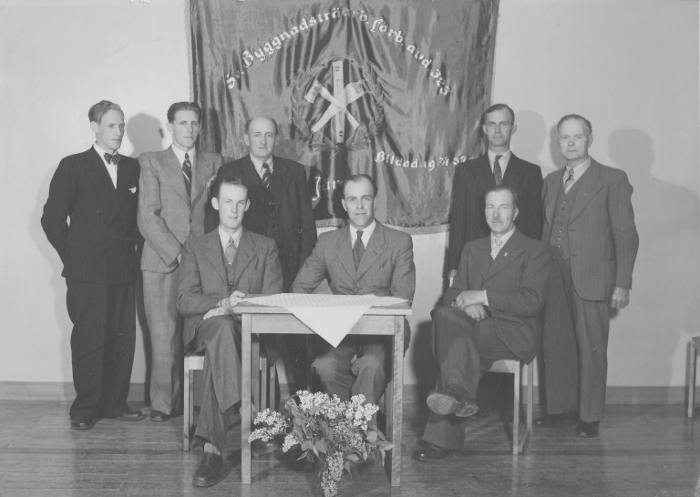 Svartvitt gruppfotografi där Emil Spring står tillsammans med andra män framför en fana i en samlingslokal.
