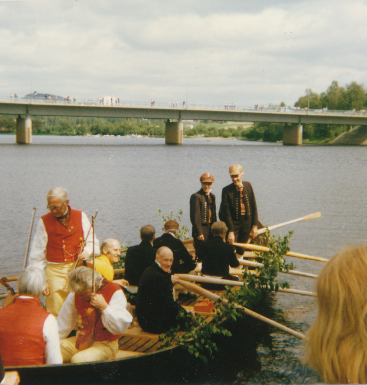 Färgfotografi av människor i båtar vid vattnet, några klädda i folkdräkt, i samband med kyrkrodd eller hembygdsarrangemang.