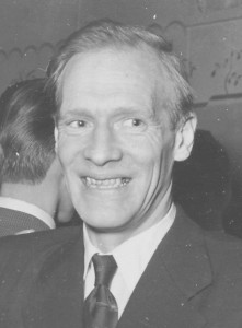 Svartvitt porträtt av Emil Leonard Spring, leende i mörk kostym och slips.