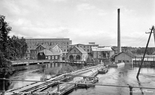 Svartvitt fotografi av industrimiljö med vatten, byggnader och skorsten vid linspinneriet i Forsa.