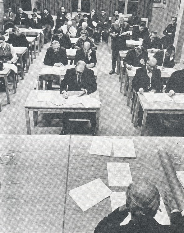 Svartvitt fotografi av mötessal med människor vid bord, sett bakifrån med en person i förgrunden.