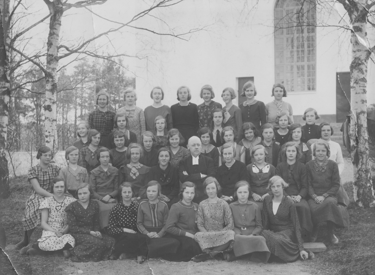 Svartvitt gruppfotografi med konfirmander och präst utomhus vid kyrkomiljö i Järvsö.