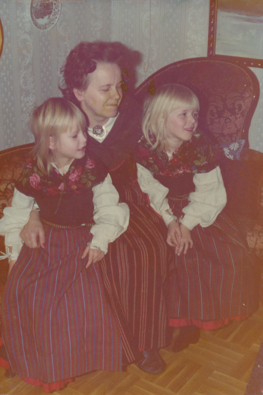 Färgfotografi av farmor Eva sittande med två barn i Järvsödräkt inomhus.