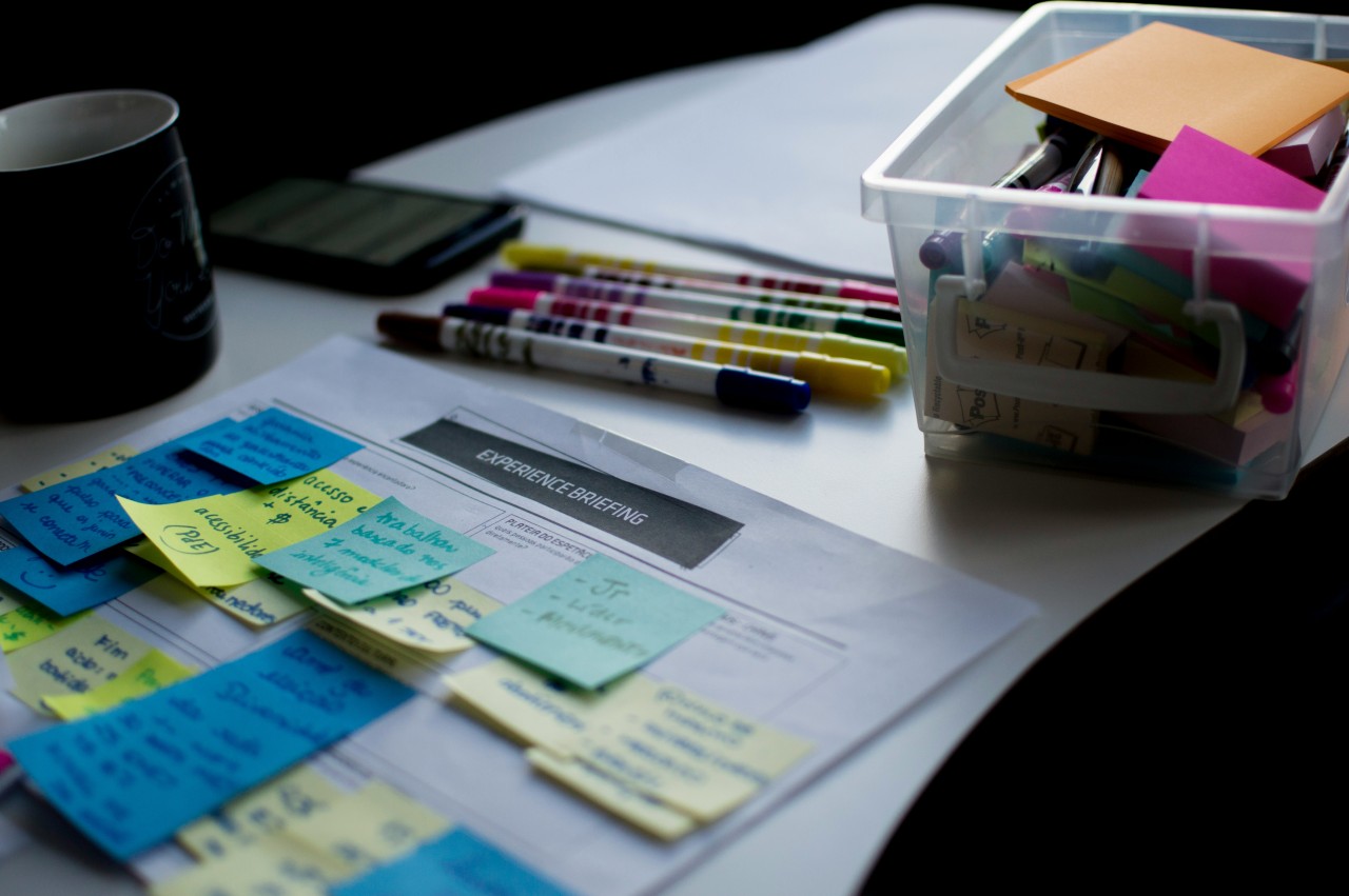 A creative workspace desk featuring design materials and planning documents. The scene includes a document titled 'EXPERIENCE BRIEFING' surrounded by colorful sticky notes in blue, yellow, and green arranged on white papers. The desk also contains colored markers, a clear plastic organizer box filled with various supplies and materials, a black coffee mug, and what appears to be a tablet or device. The setup suggests an active design thinking or user experience planning session with organized brainstorming materials.