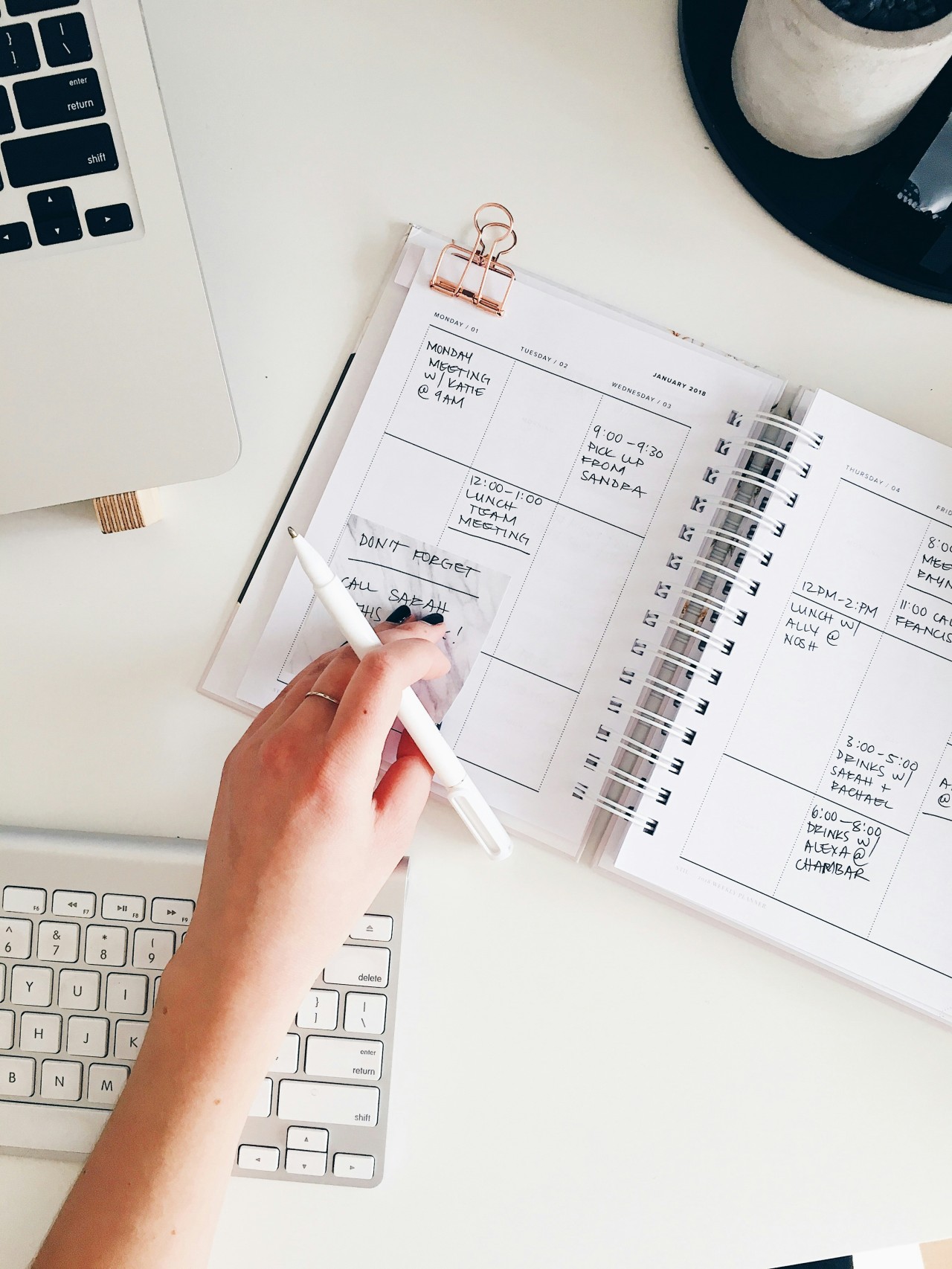 An overhead view of a hand holding a white pen over an open weekly planner filled with handwritten appointments, next to a laptop keyboard on a white desk