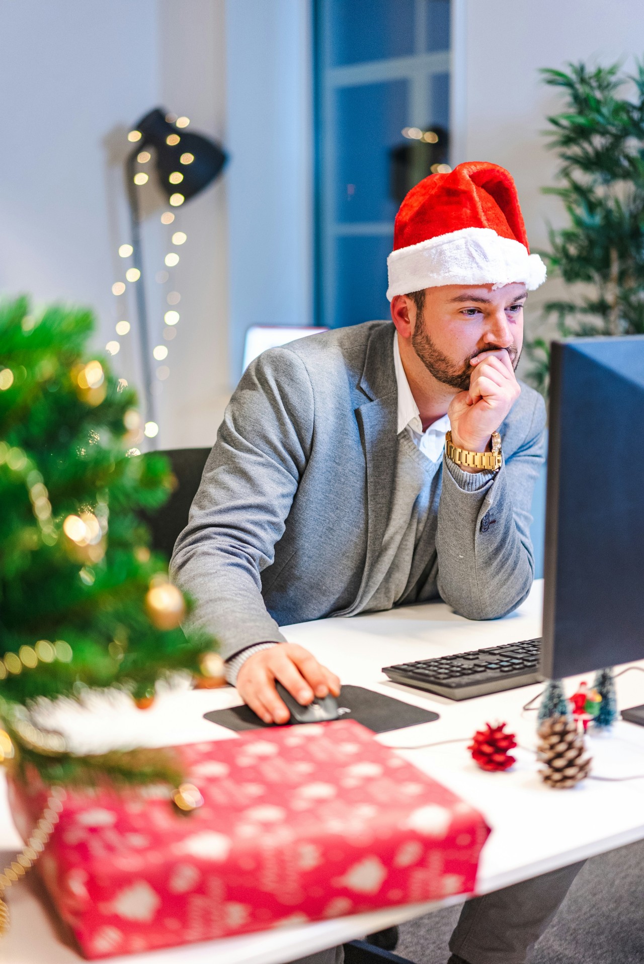 A businessman wearing a red Santa hat and gray suit sits at a white desk working on a laptop in a modern office decorated for Christmas, with a lit Christmas tree, wrapped red gift boxes, pinecones, and string lights creating a festive holiday workspace atmosphere