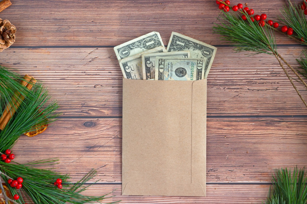 A brown paper envelope containing several US dollar bills sits on a rustic wooden surface, surrounded by festive holiday decorations including pine branches with red berries and pinecones, creating a seasonal gift-giving scene