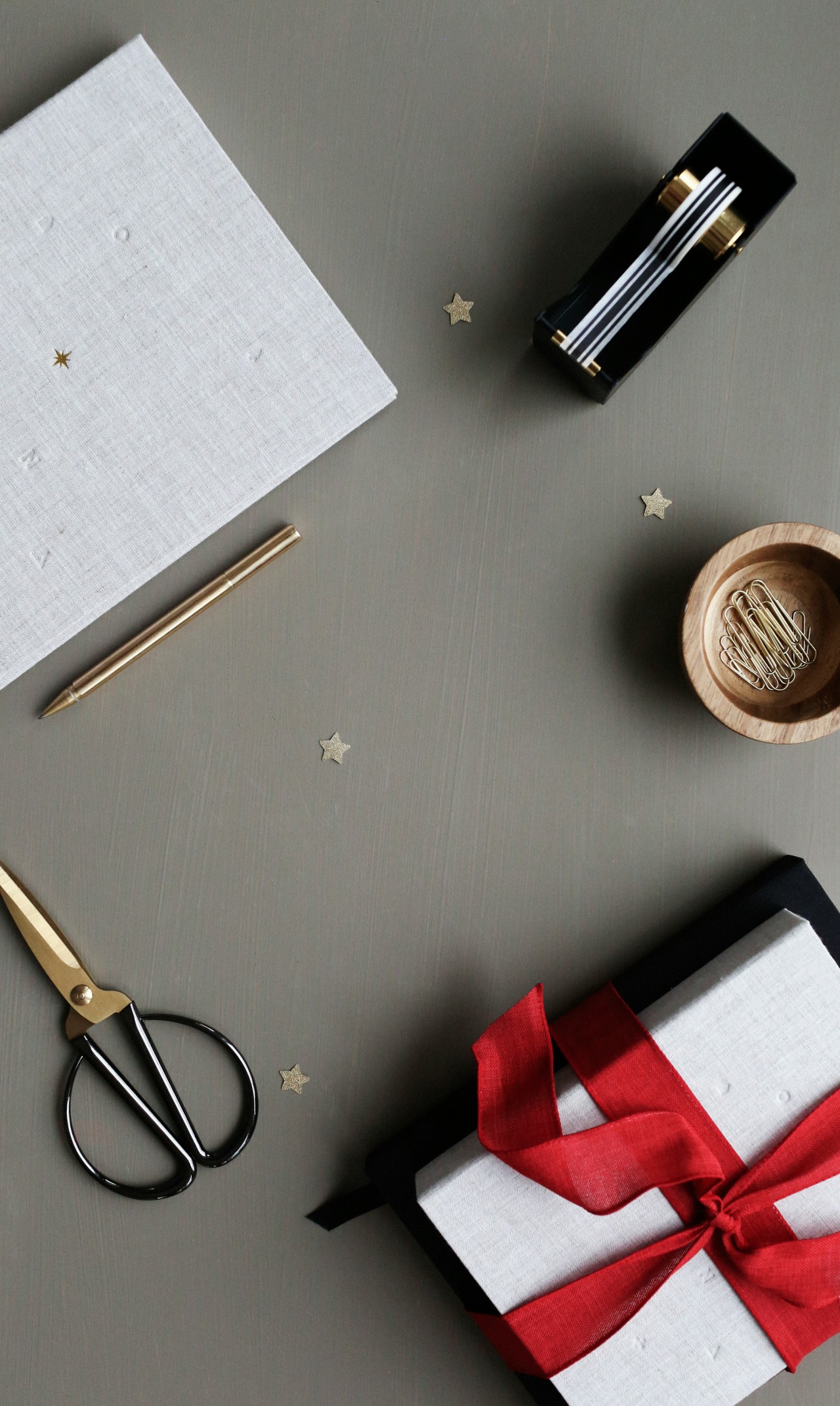A flat lay arrangement of gift wrapping and stationery items on a gray surface. The scene includes white textured paper with small gold star patterns, a gold pen, black scissors with gold handles, a black pen case or holder containing white and gold pens, a wooden bowl filled with gold paper clips, small gold star decorations scattered around, and a white gift box tied with a red satin ribbon. The composition has a minimalist, elegant aesthetic with a neutral color palette accented by gold and red details.