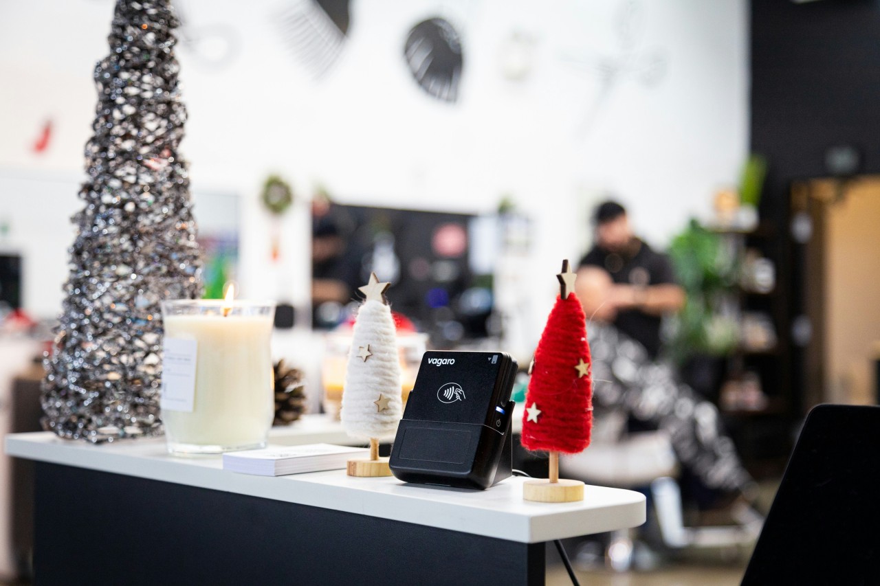 A modern business counter decorated for Christmas featuring a black Vagaro payment terminal with contactless payment capability, flanked by small decorative Christmas trees in white and red felt with gold stars, a lit candle, pinecones, and a sparkly silver Christmas tree, with blurred office workspace in the background
