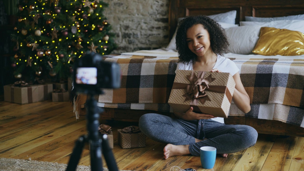 A young woman with curly dark hair sits cross-legged on a wooden floor, smiling while holding a wrapped gift box with polka dot paper and a brown ribbon. She's wearing a white t-shirt and jeans, positioned in front of a couch with a brown and white plaid throw blanket. A smartphone on a tripod is recording her, with its screen visible showing her image. Behind her is a Christmas tree decorated with warm white lights and ornaments, and wrapped presents are visible beneath it. A blue mug sits on the floor nearby. The setting appears to be a cozy living room with exposed brick walls.