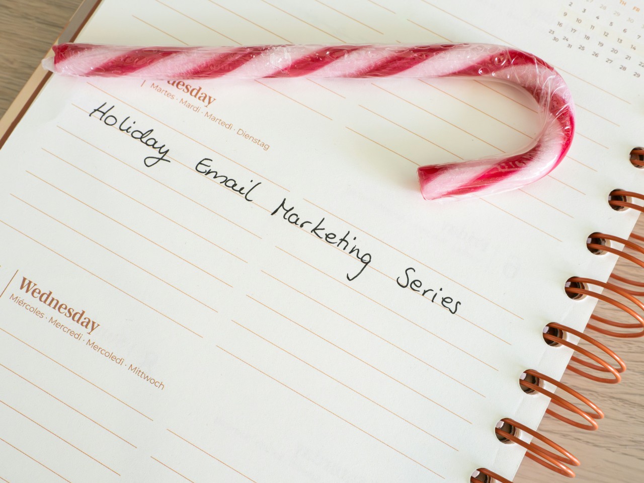 A spiral-bound planner or calendar page showing Tuesday and Wednesday entries. On Tuesday's page, handwritten text reads 'Holiday Email Marketing Series' in cursive script. A red and white striped candy cane is placed diagonally across the top of the page. The planner has copper-colored spiral binding on the right side and sits on a light wooden surface.