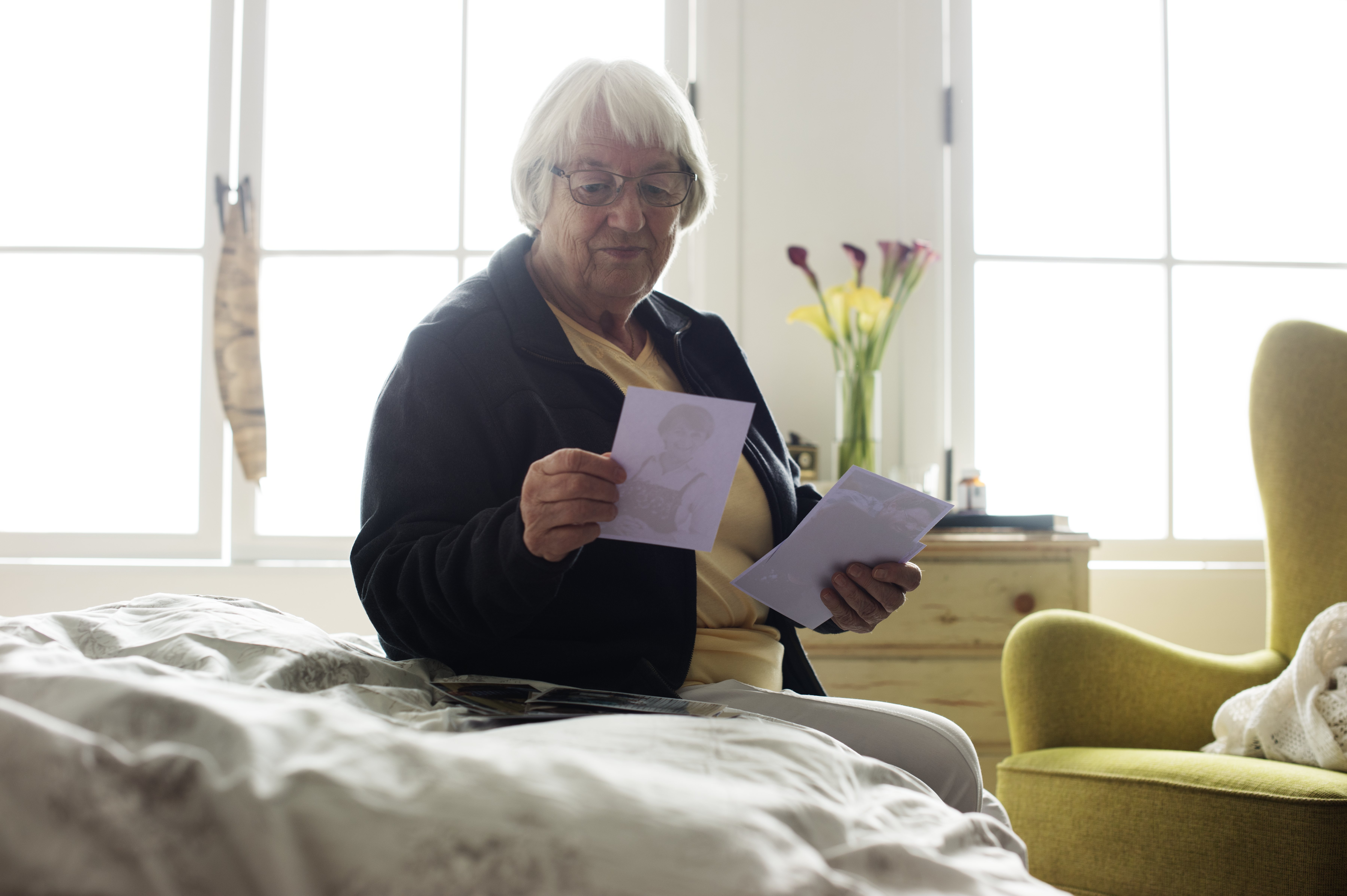 An older woman with white hair reads a handwritten letter while sitting on the edge of her bed in a cozy, sunlit room.