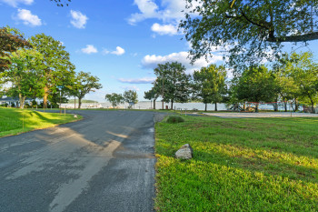 Paved road with adjacent green lawn, surrounded by mature trees, and waterfront access visible in the background