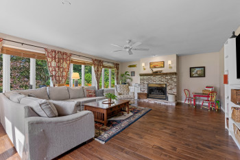 Spacious living area featuring wood-finish flooring, a brick fireplace with a mantle, and large windows with horizontal blinds