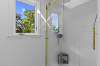 Shower enclosure featuring a clear glass door with brass-finish framing, white ceramic tile walls, a decorative tile band, a skylight, and a fixed showerhead with a brass-finish grab bar