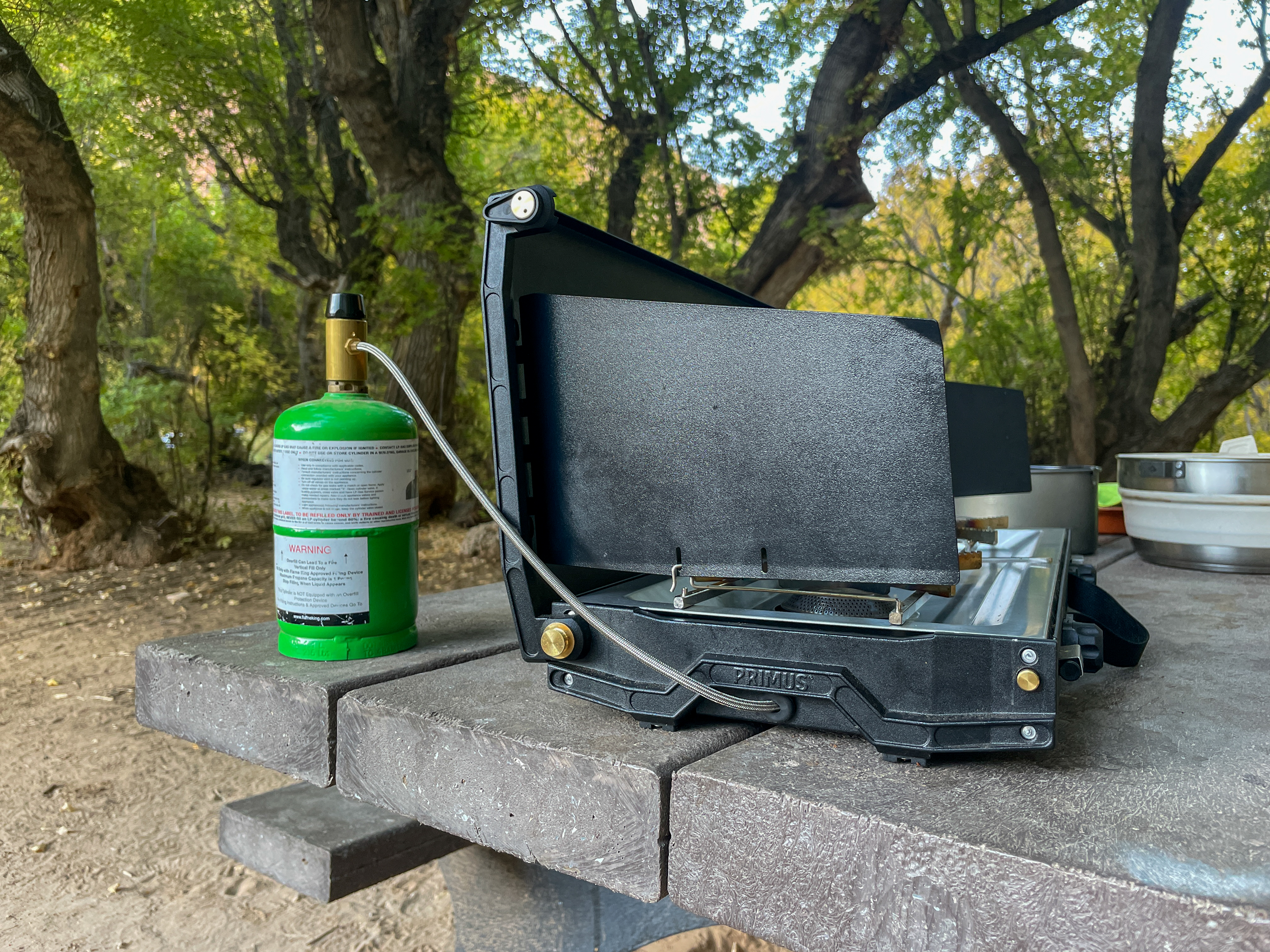 the primus alika stove set up on a picnic table with the flexible gas hose connecting to a green propane canister