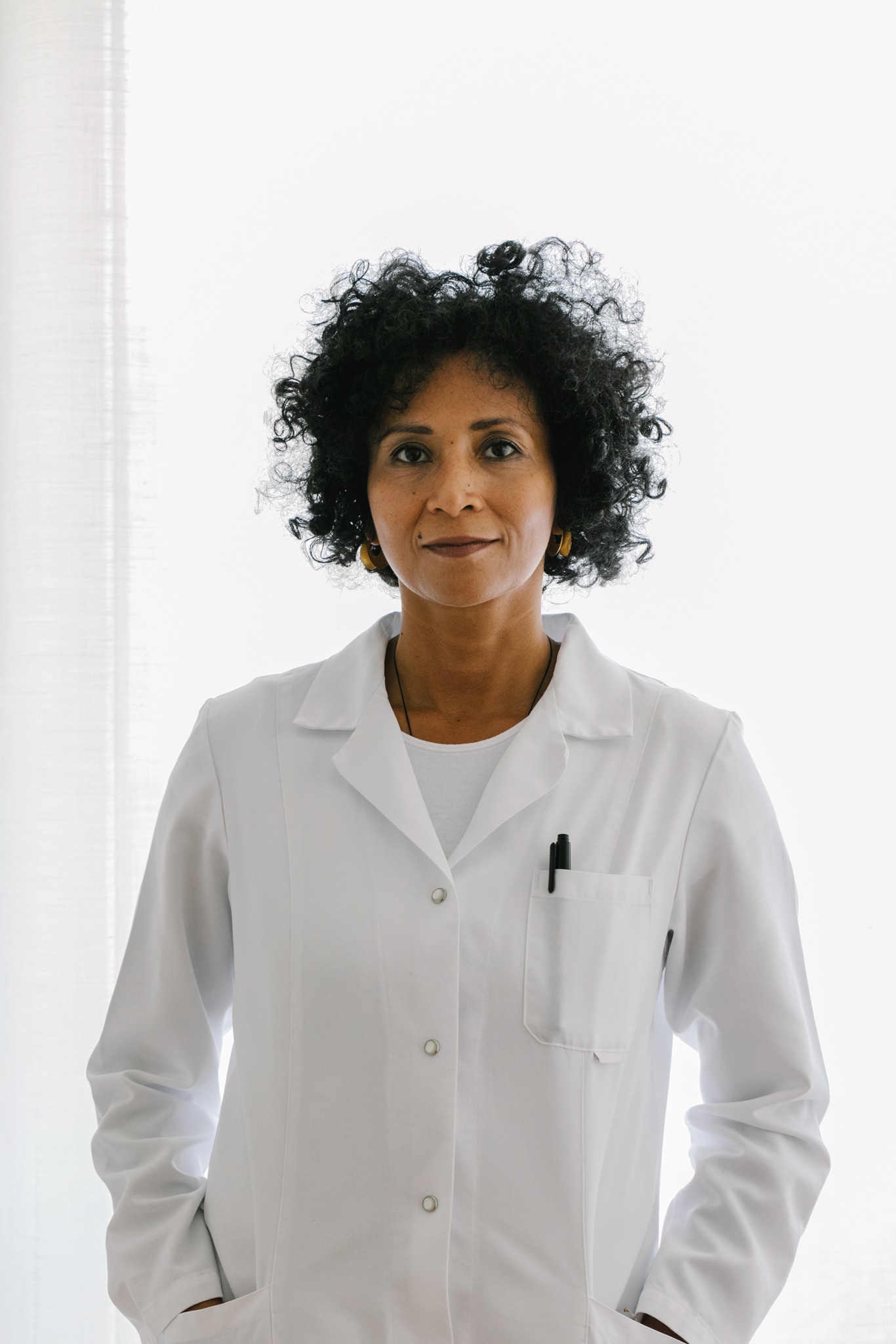 Indoor portrait of a female physican of hispanic ethnicity  She is wearing a white lab coat and is standing in front of a large window 