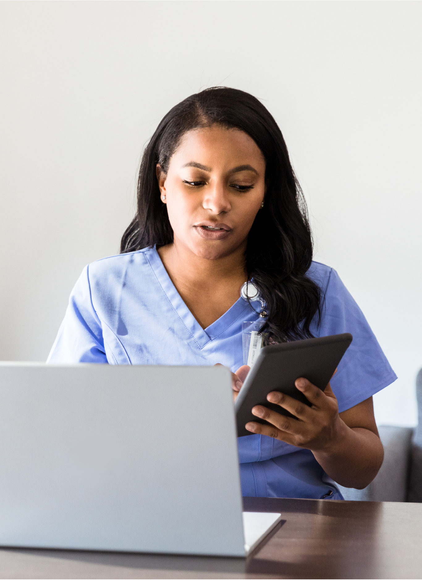 A serious doctor checks her appointment calendar while working in her office  She is using a digital tablet to check her calendar  She is sitting in front of an open laptop 