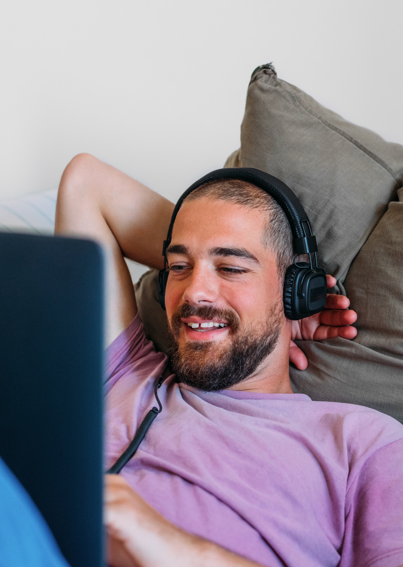 An attractive young man lying on the bed, wearing headphones, using a laptop, smiling