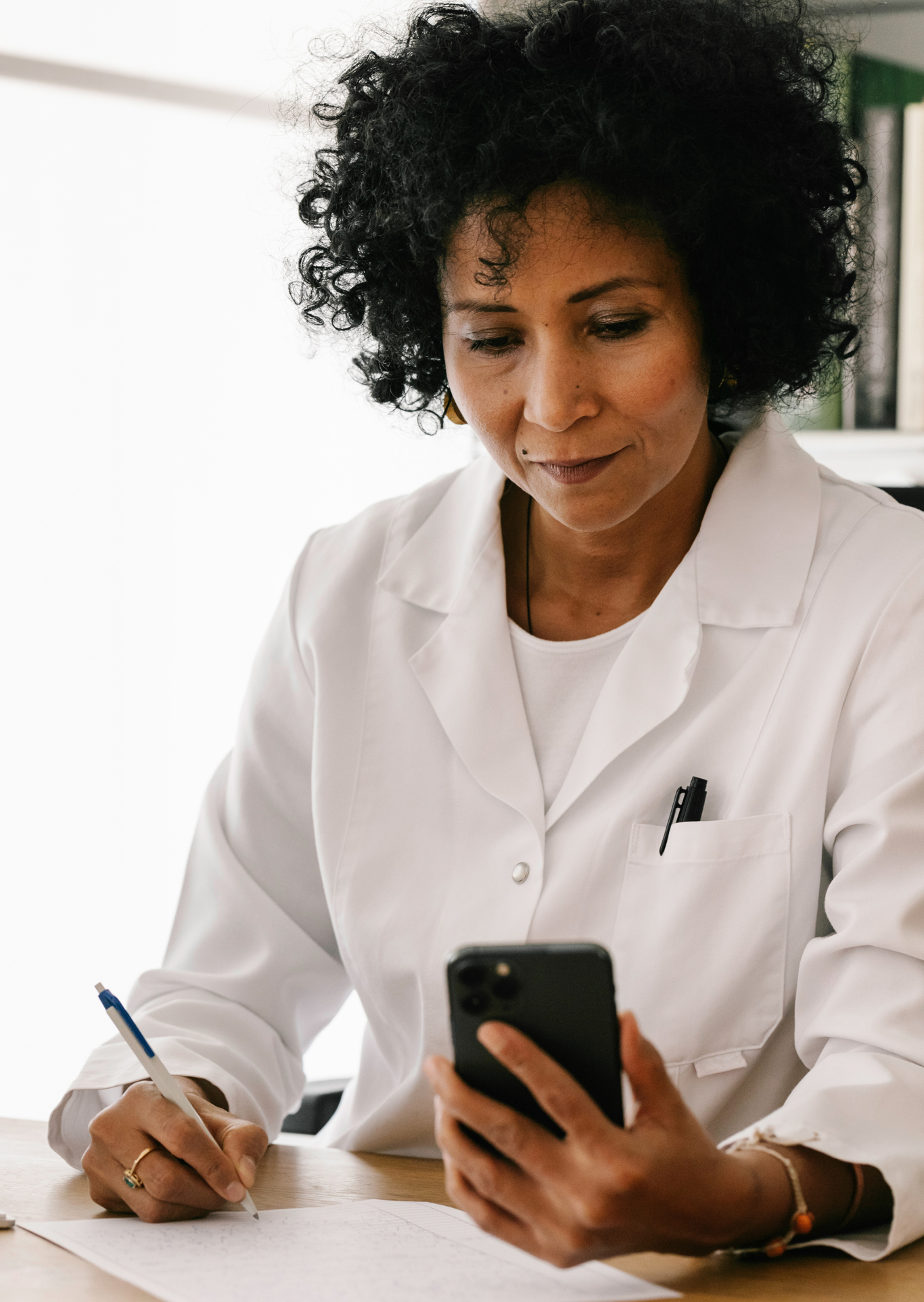 A latin woman doctor talking to a patient via video  She is holding a cellphone in her left hand and is writing down observations simultaneously  
