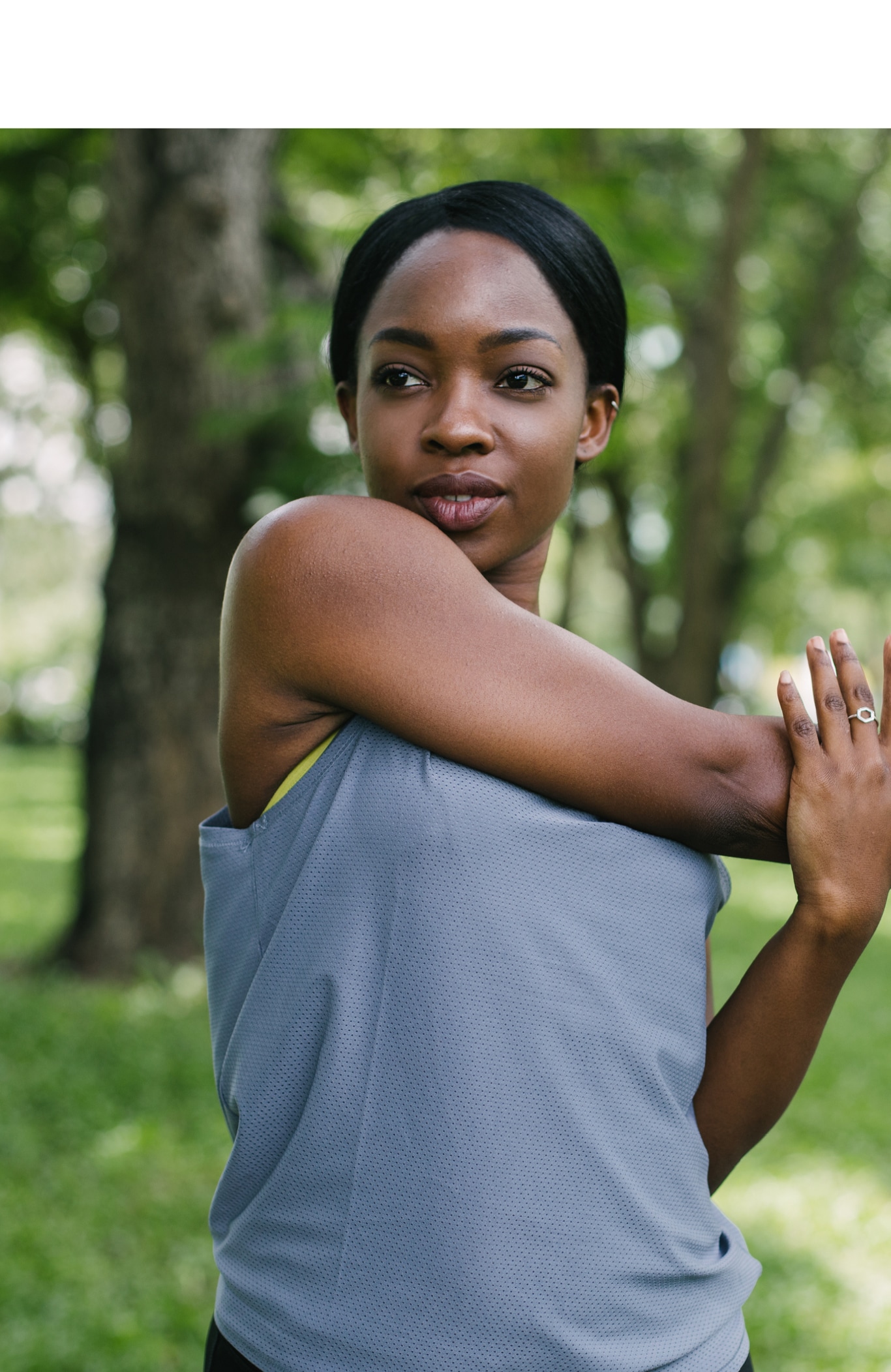 A young black woman doing a warm up stretch in the park on the road amidst green grass and trees 