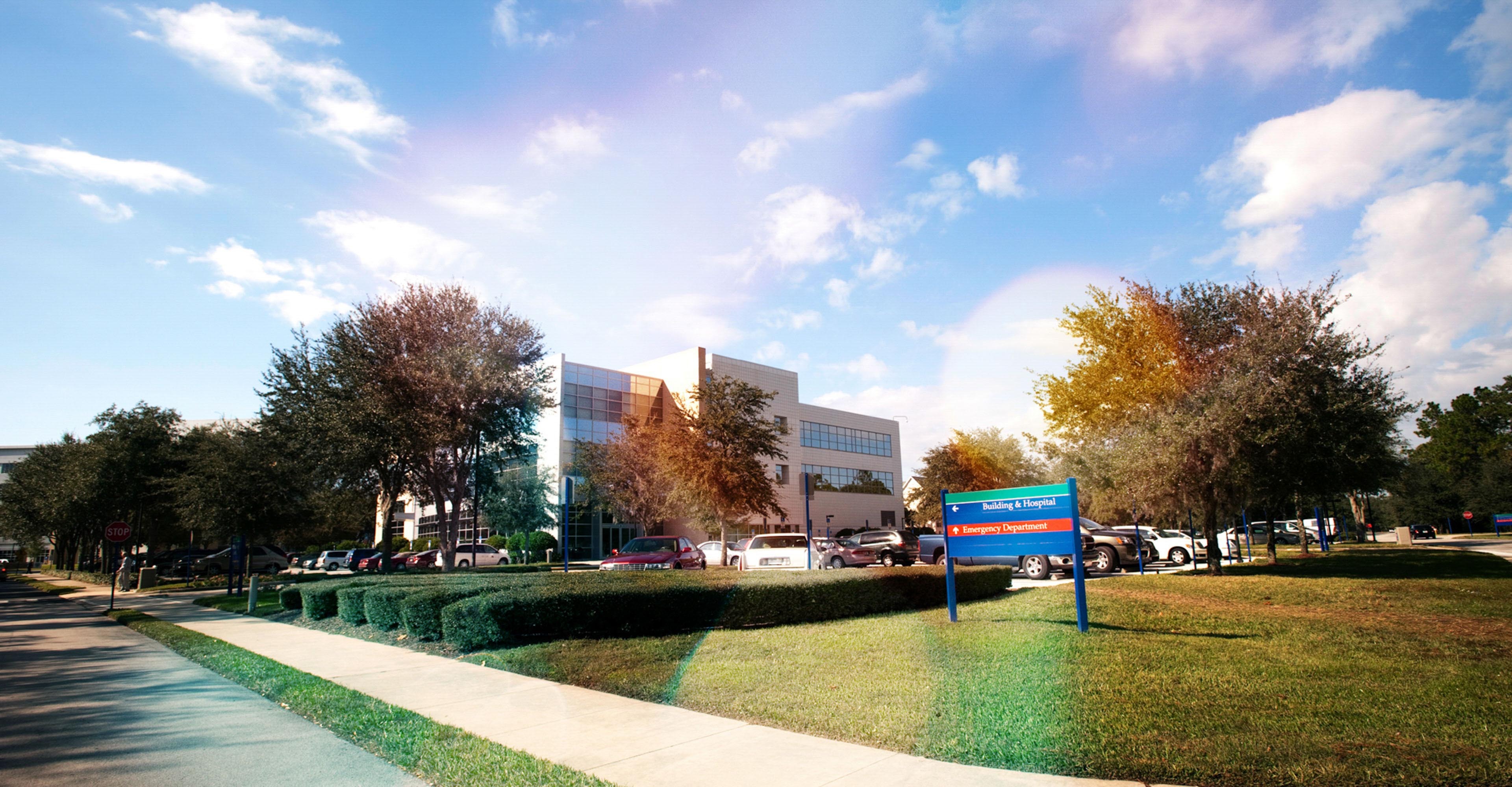 Modern building and parking lot under a partly cloudy sky. Lush trees surround the area.