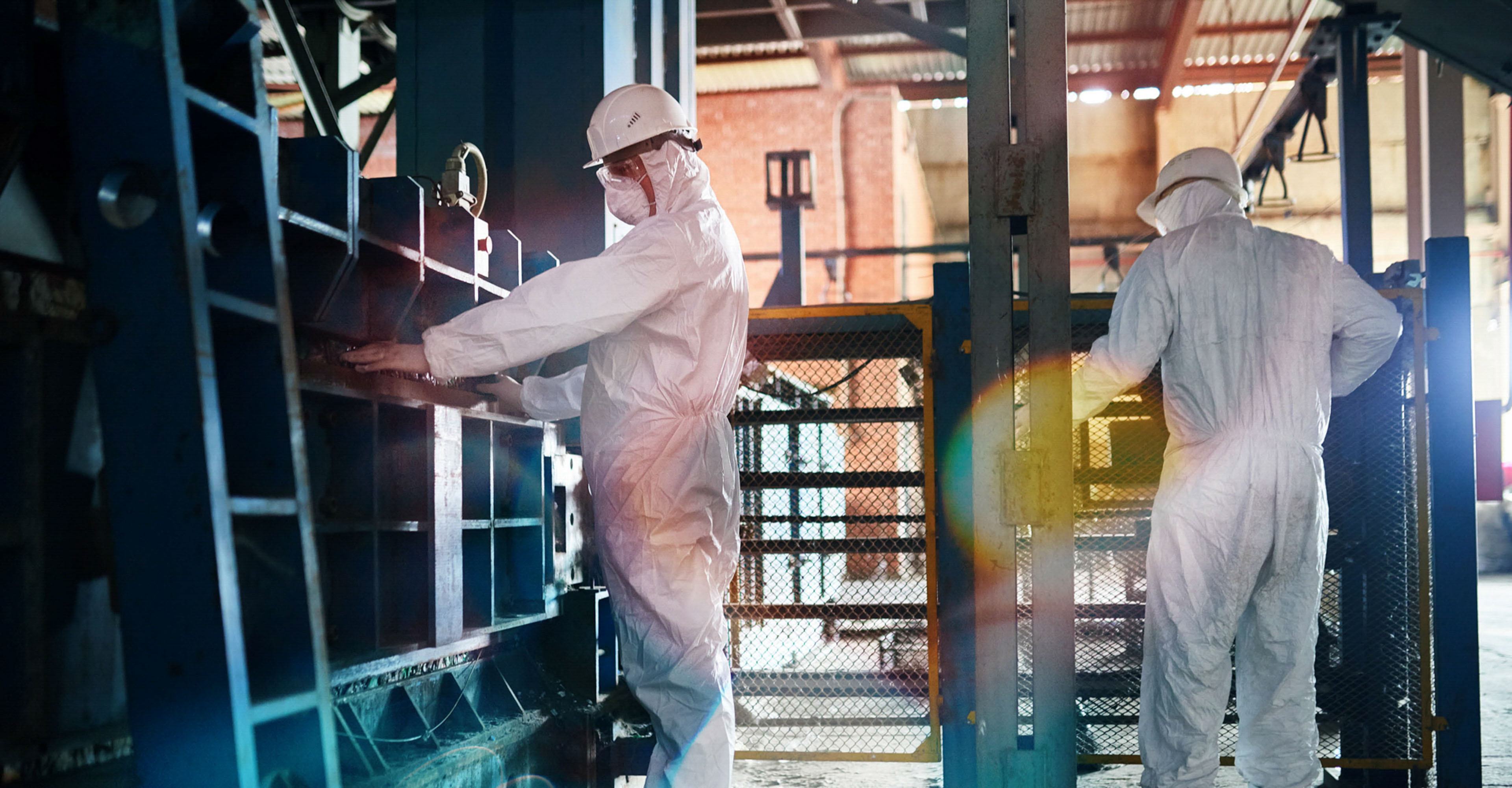 Two workers wearing biohazard suit and hardhats working in open workshop of industrial plant