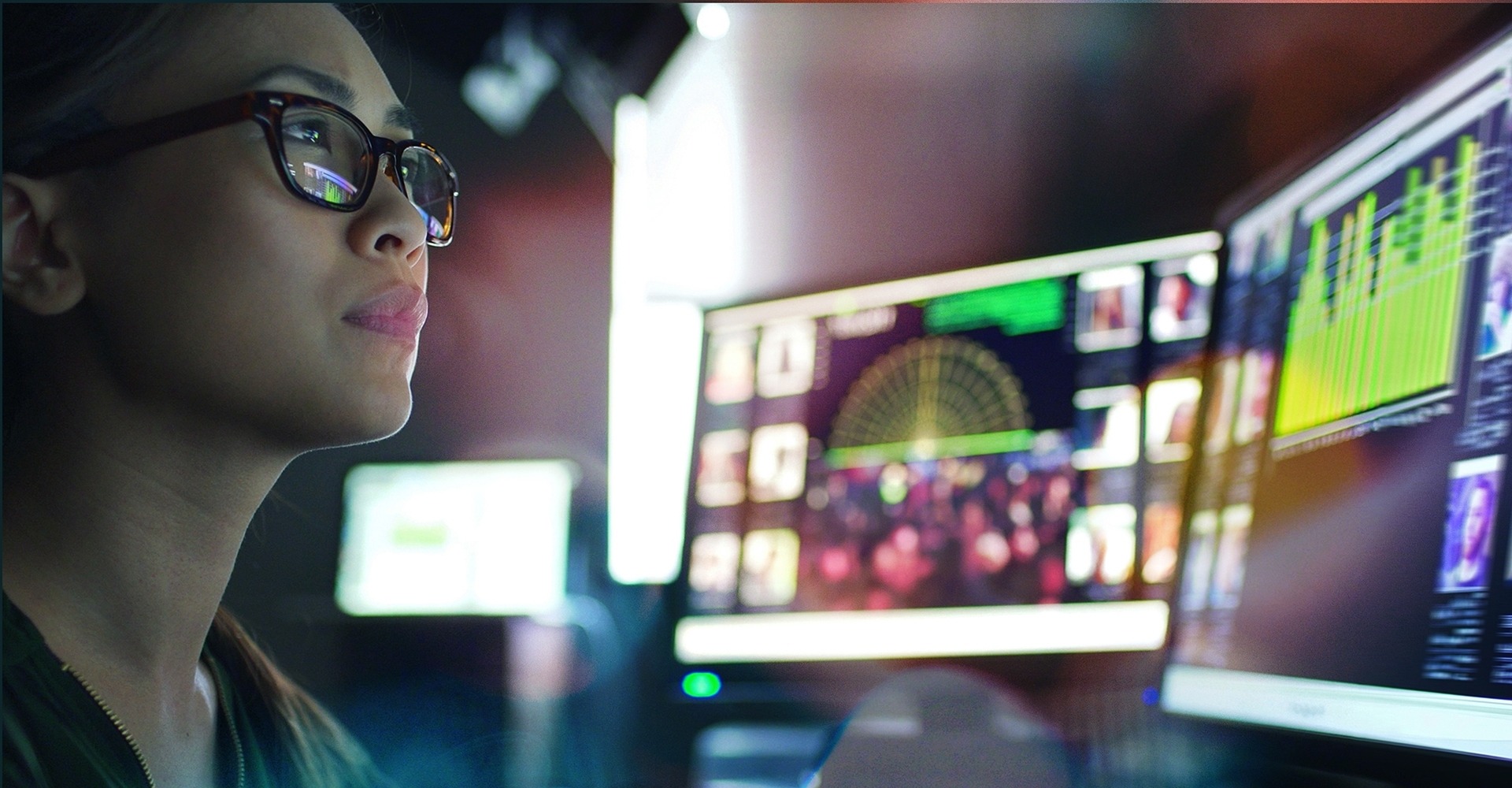 A young asian woman sitting down at her desk where she’s surrounded by 3 large computer monitors