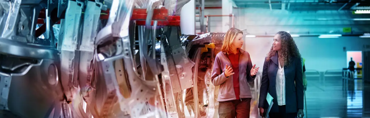 Female engineers discussing by car chassis walking on an aisle in a factory. 