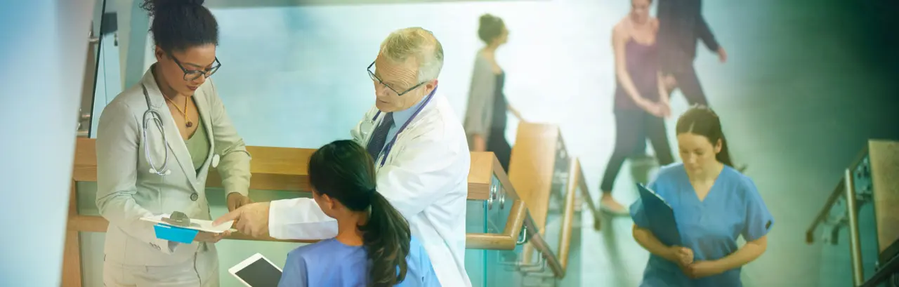 Medical professionals stand on a stairwell of a modern hospital and discuss some case notes.