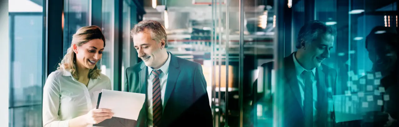 Two collogues walking in an office building discussing something on the paper the woman is holding