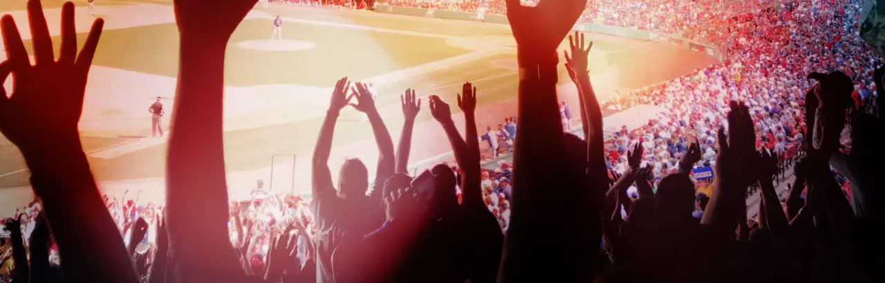 Image of fans at a baseball stadium, focus on some fans standing up with their hands in the air.